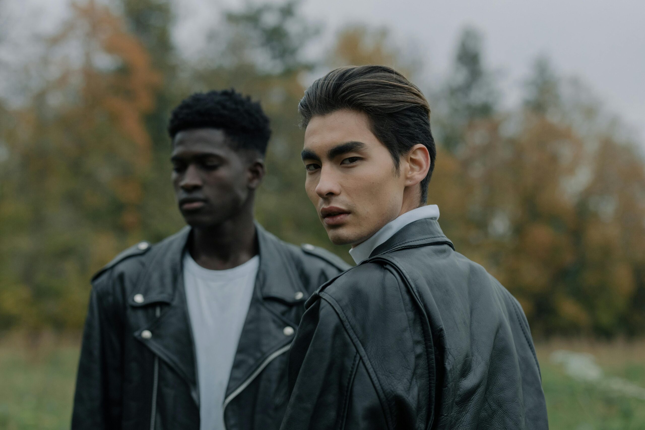 Two stylish men wearing leather jackets outdoors in an autumn setting.